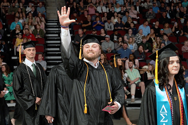 A smiling man in a cap and gown is shown waving with his arm outstretched.