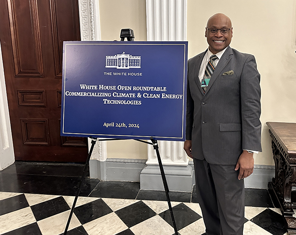 A man in a grey suit stands next to a sign on a display that says "White House Open Roundtable Commercializing Climate and Clean Energy Technologies; April 24th, 2024."