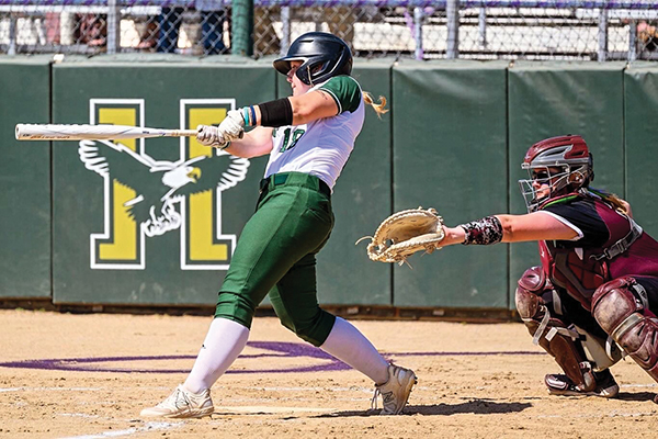 A woman wearing a green and white Husson University softball uniform is at the plate with a bat extended. A catcher with a mitt extended in a maroon uniform crouches behind her.