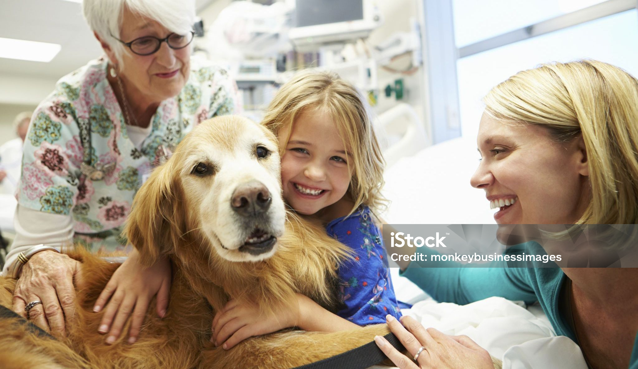 Young patient sits on a bed with a dog while a woman and healthcare worker smile and look on.