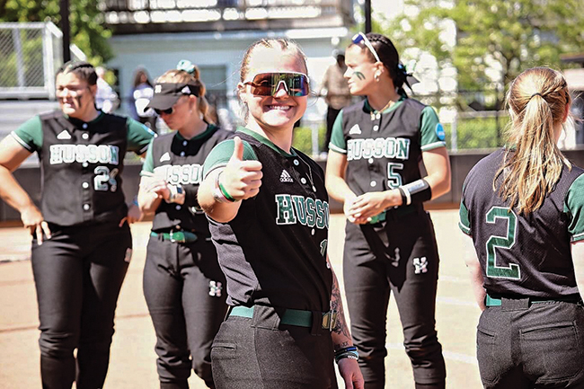 A young woman wearing sunglasses and wearing a black and green Husson University softball uniform gives a thumbs up to the camera. Other young women in the same uniform stand around her.