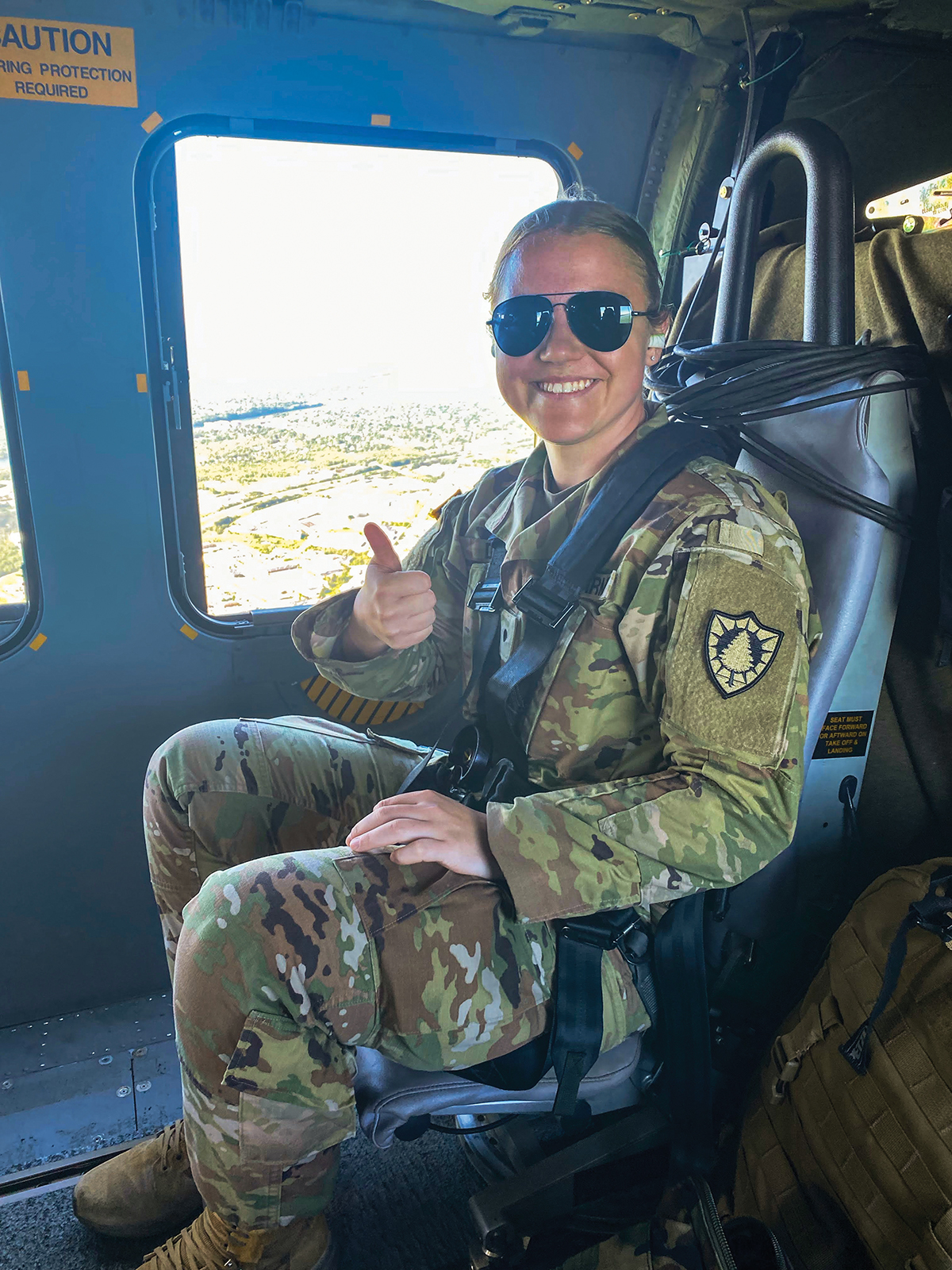 A young woman wearing sunglasses and green fatigues is strapped into an aircraft seat smiling and giving a thumbs up to the camera.