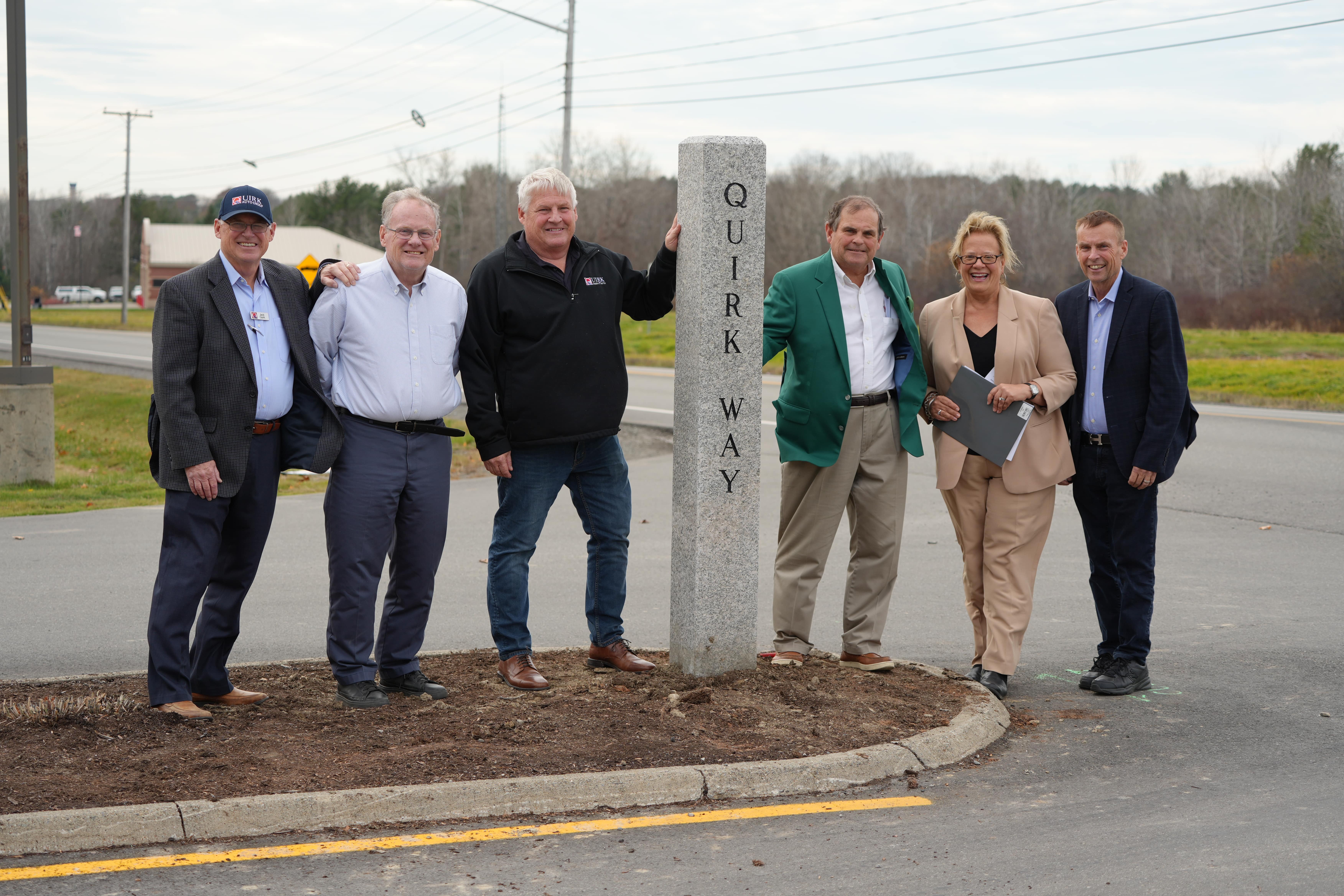 Members of the Quirk family and Husson University community stand next to the granite post marking the newly named entrance to Husson Univesity. The post says "Quirk Way."