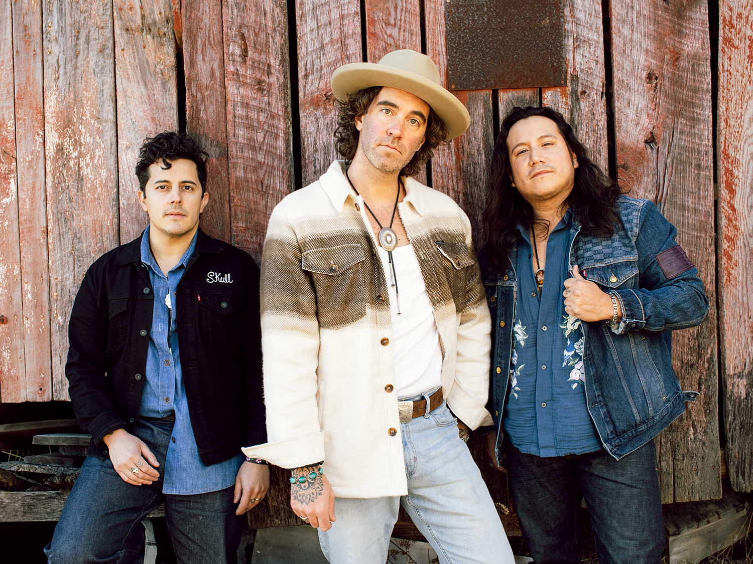 Three men look at the camera. They are shown from the thigh up and have the wall of a red barn behind them.