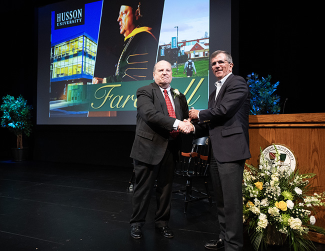 Two men are shown on a stage, shaking hands, with a video screen behind them with the word "Farewell" on it.