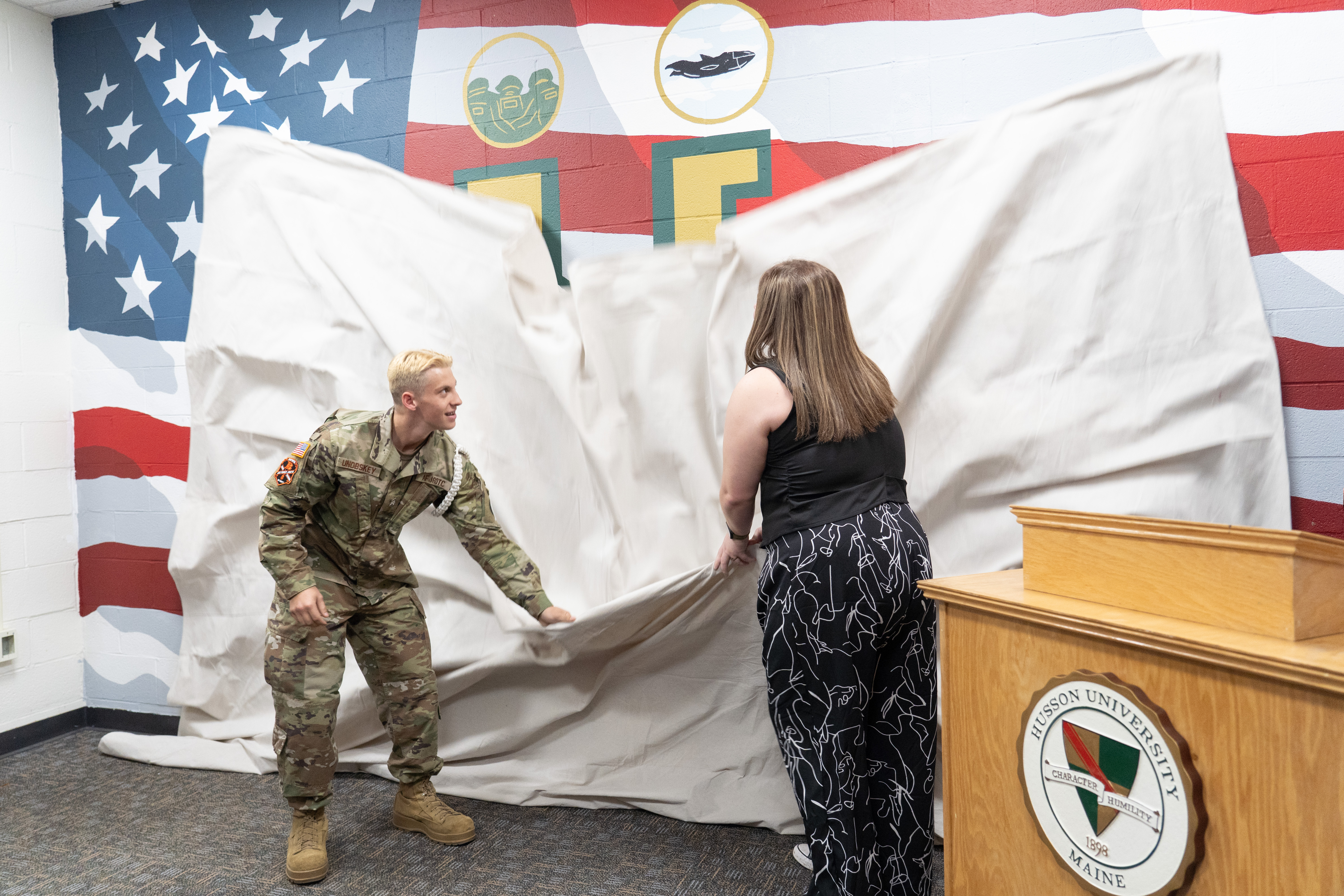 Two young people pull a cloth down, revealing a painted mural on a wall. The cloth is partially obscuring the wall as it falls.