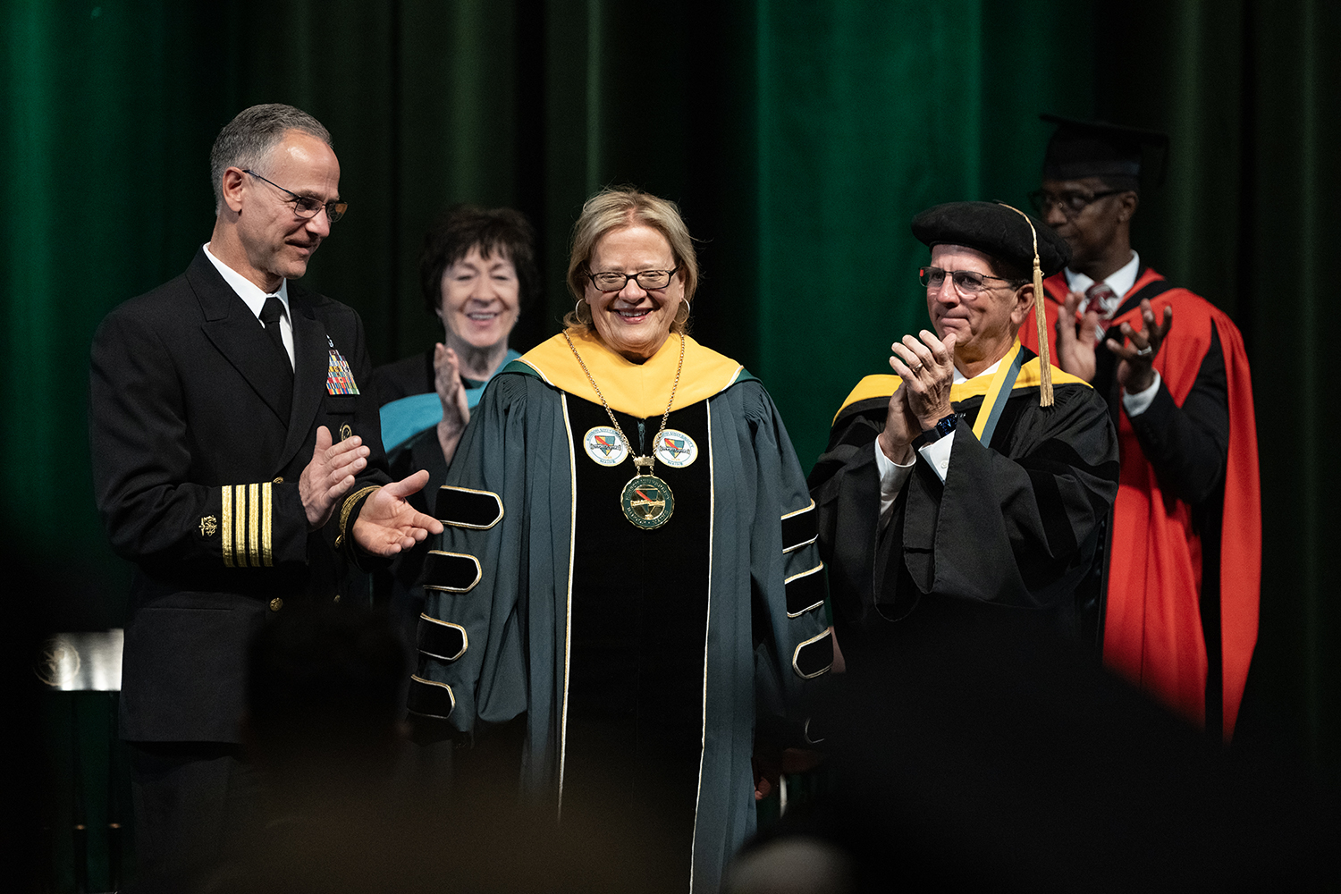 A smiling woman in regalia stands next to two smiling and clapping men on a stage.