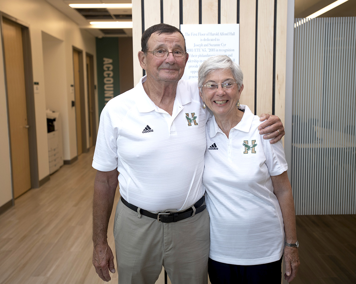 A man and woman wearing white Husson shirts stand together in this photo. The man has his arm around the woman.