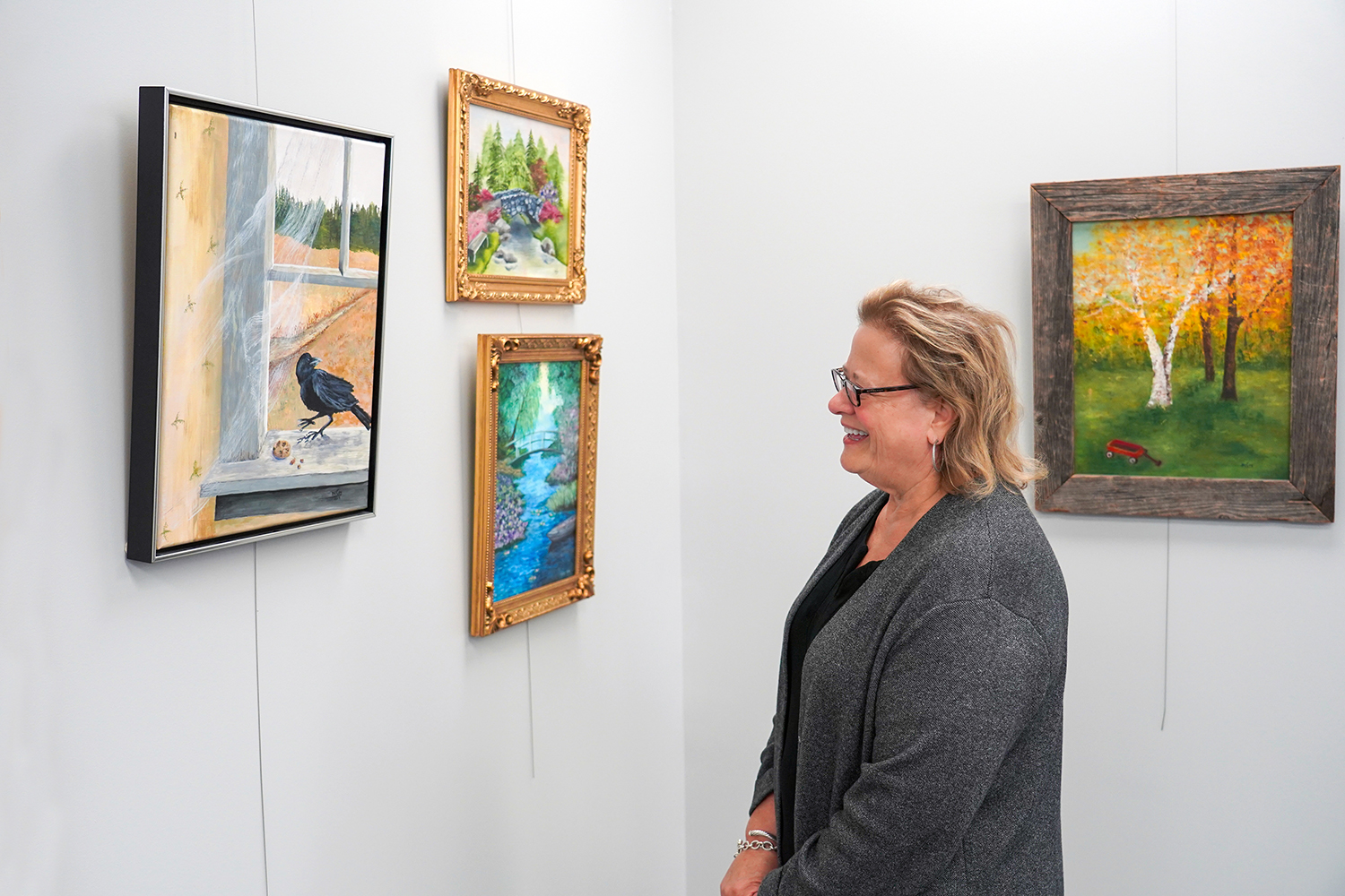 A blond woman smiles at a painting hung on a wall in a gallery.