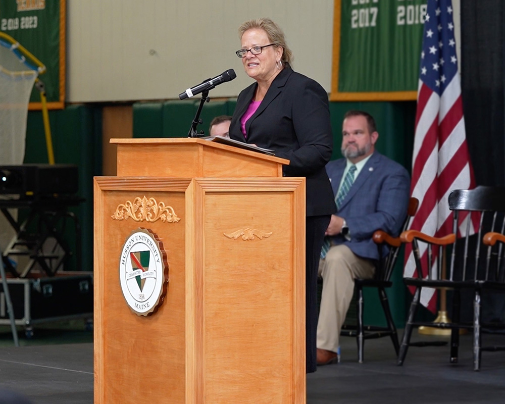 A blond woman in a black suit with a pink shirt stands at a Husson University podium