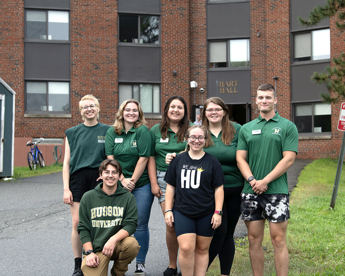 A group of young people wearing Husson University gear stand in a group shot in front of a brick building with the words Hart Hall. They are smiling.