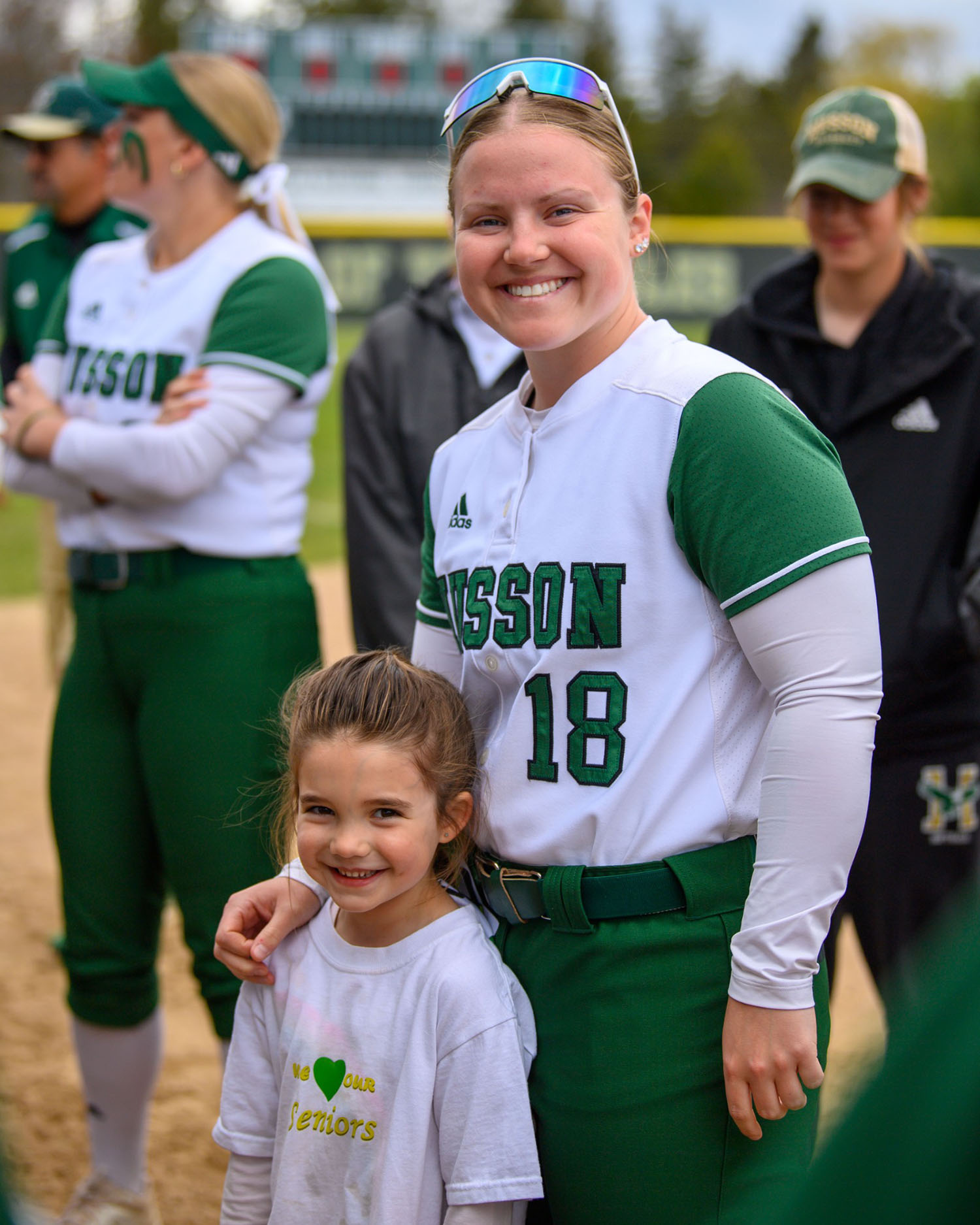 A smiling woman in a green and white Husson softball uniform is standing with other players in the background and a little girl in front of her.