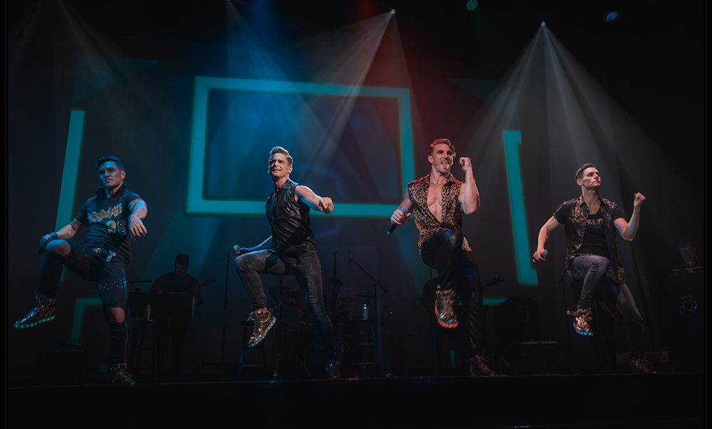 Four men in black stand on a stage singing and dancing.