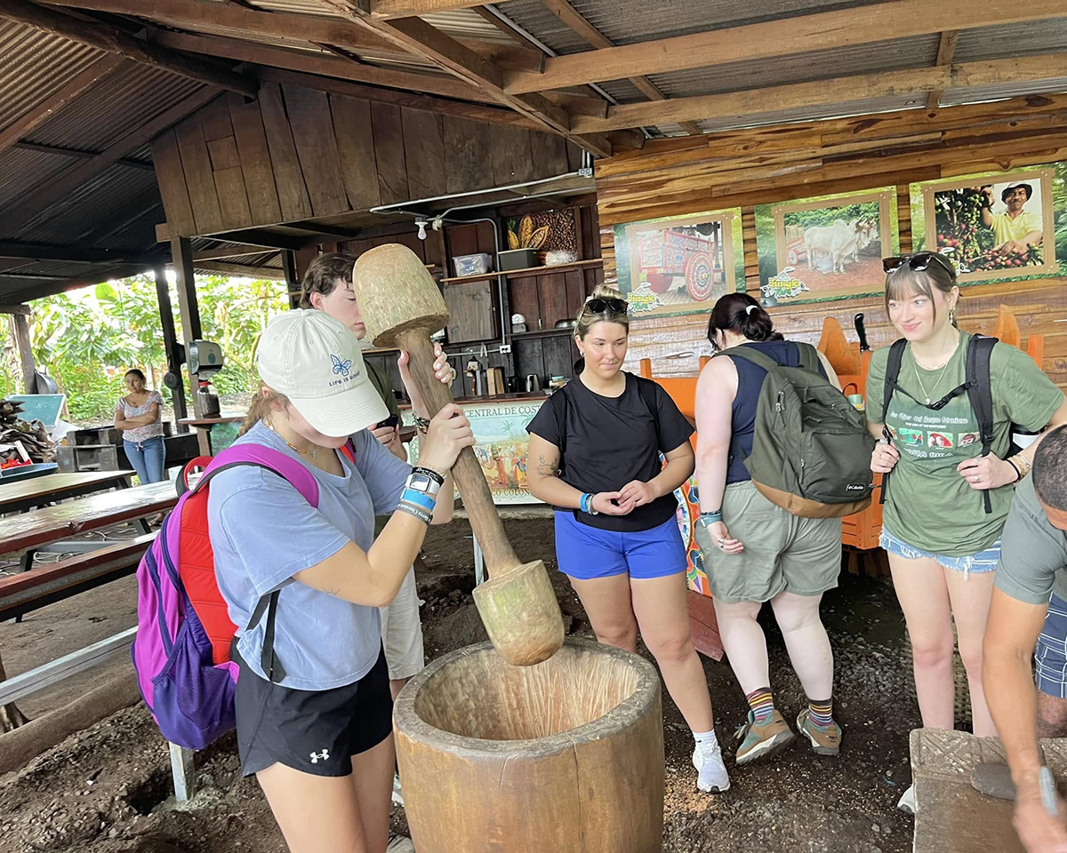 A young woman uses a large wooden tool that's plunged into a barrel. Others look on.