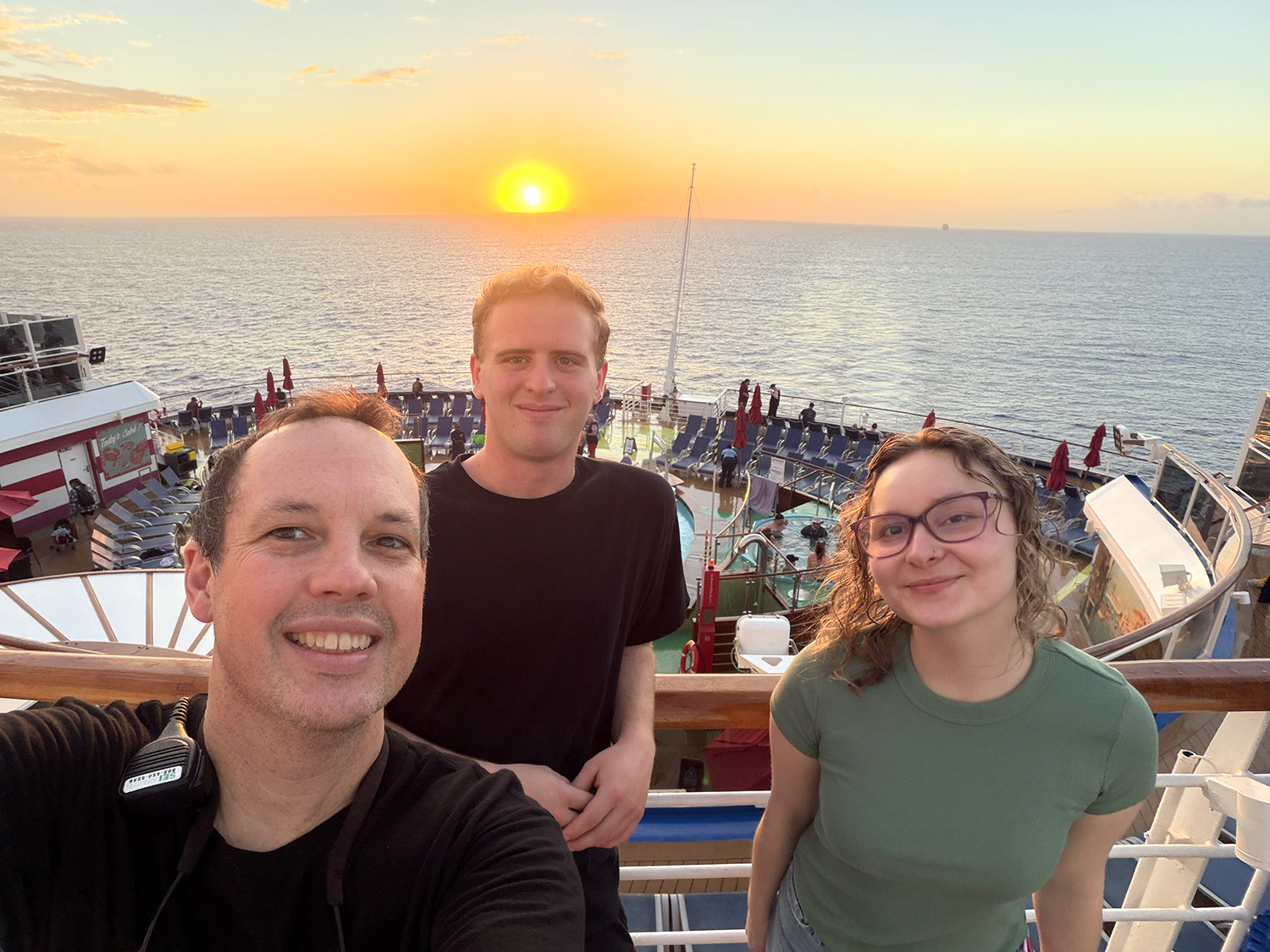 Three people smile at the camera. They are standing on a cruise ship with a sunset behind them.