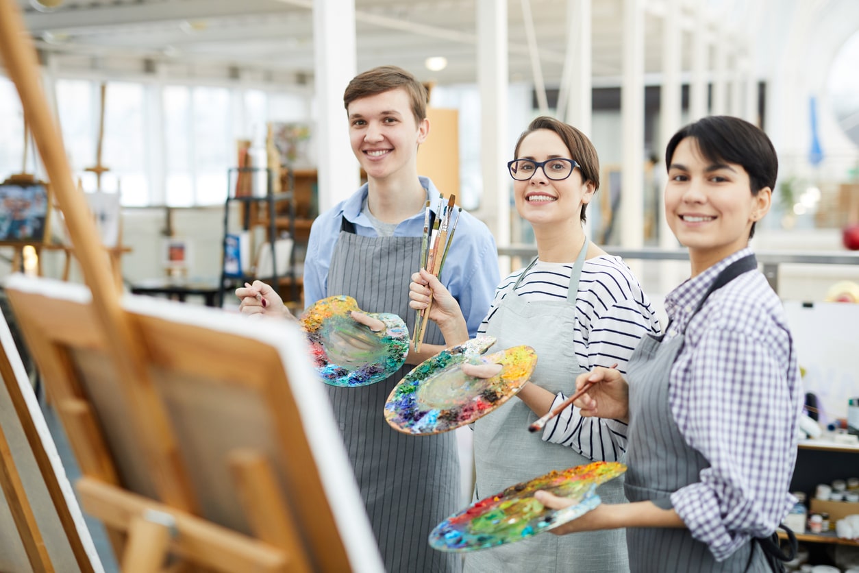 Three artists holding painting supplies while smiling at the camera.