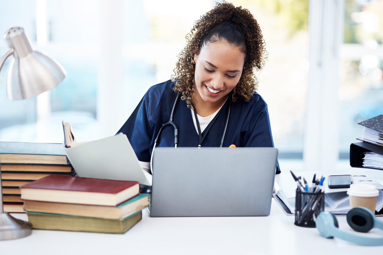 Nurse working on a laptop that is surrounded by stacks of books.